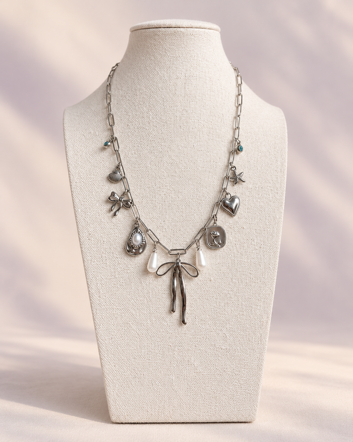 Silver paperclip charm necklace with bows, pearls, heart, shell, and starfish on a cream linen display.