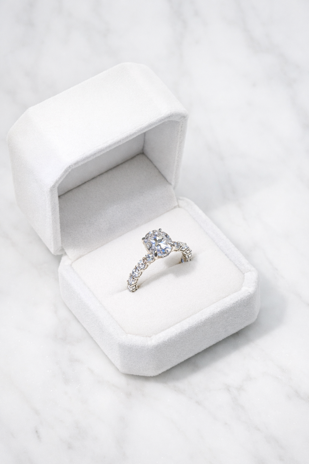 Diamond engagement ring in a white velvet ring box on a pale stone surface.