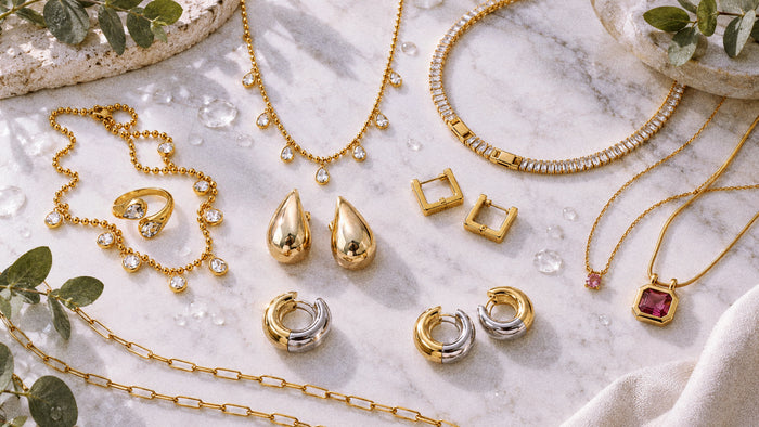 Gold and mixed-metal waterproof jewelry styled on pale stone with water droplets and eucalyptus.