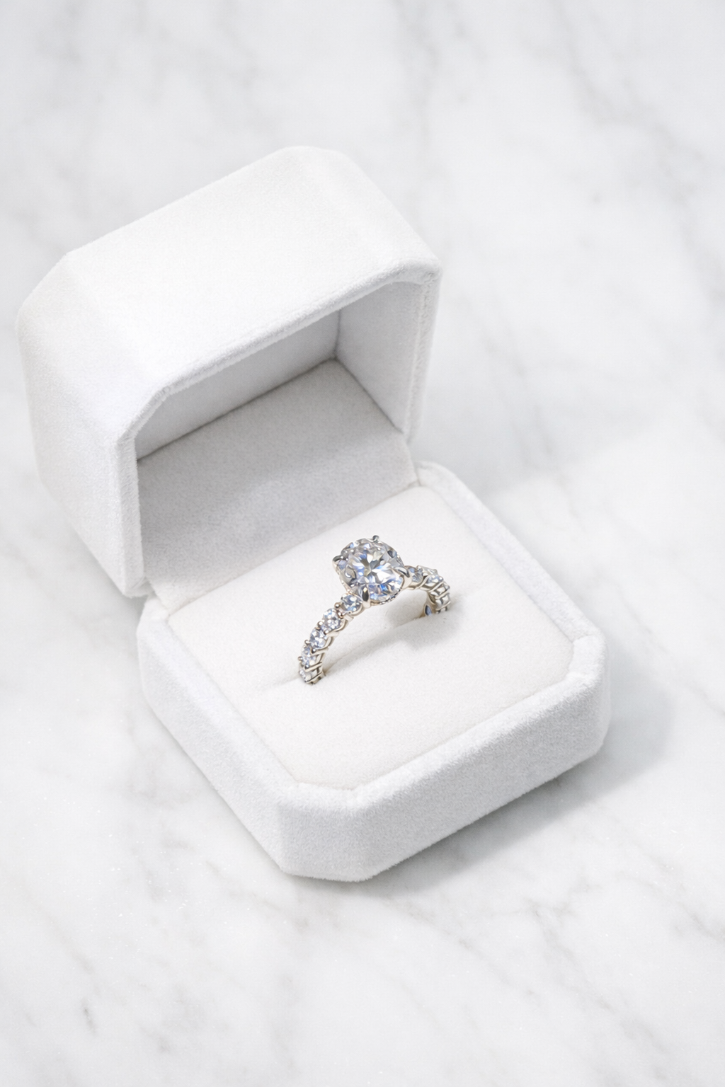 Diamond engagement ring in a white velvet ring box on a pale stone surface.