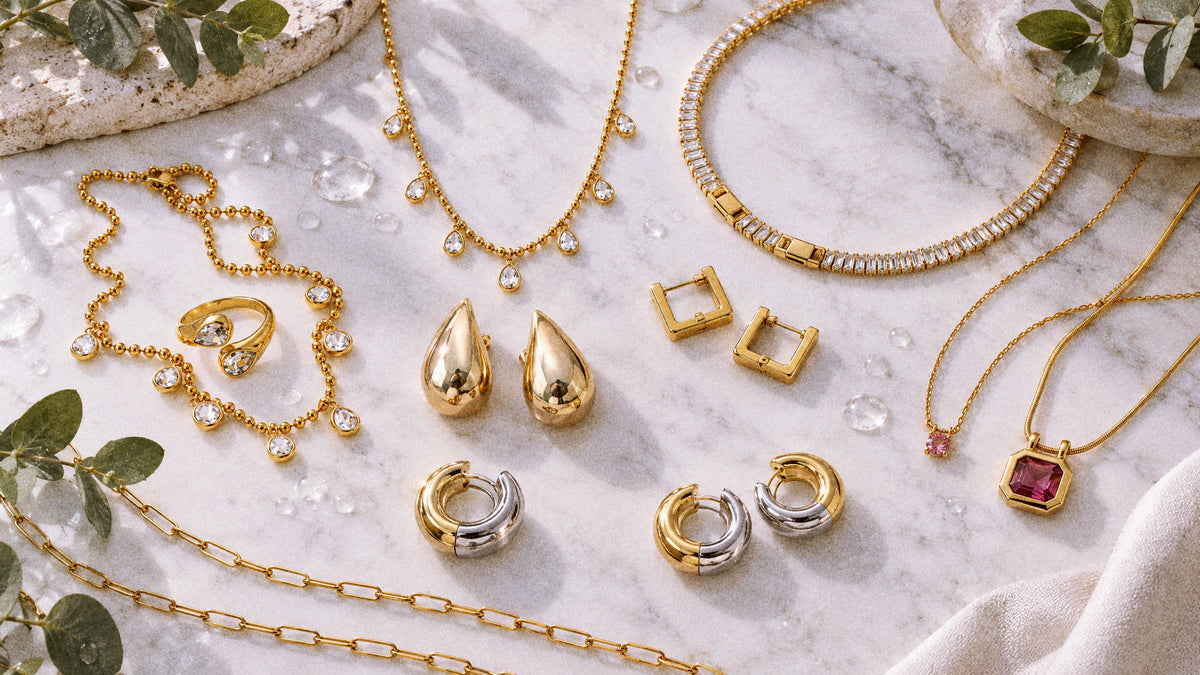 Gold and mixed-metal waterproof jewelry styled on pale stone with water droplets and eucalyptus.