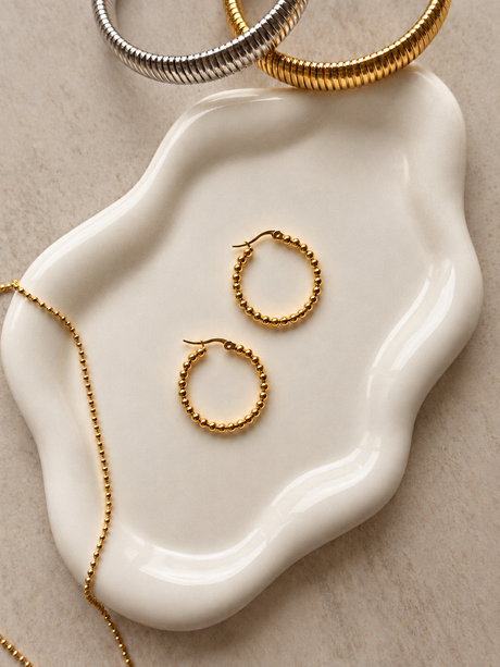 Pair of gold bubble hoops on a white ceramic dish; other jewelry is styling only